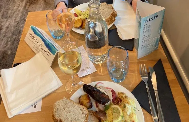 Wooden table with two plates of food, two glasses of white wine, cutlery, and water.