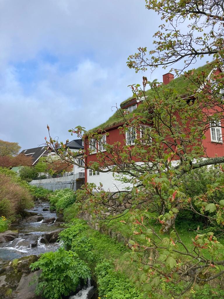 Red house with grass on top and greenery around overlooking stream.
