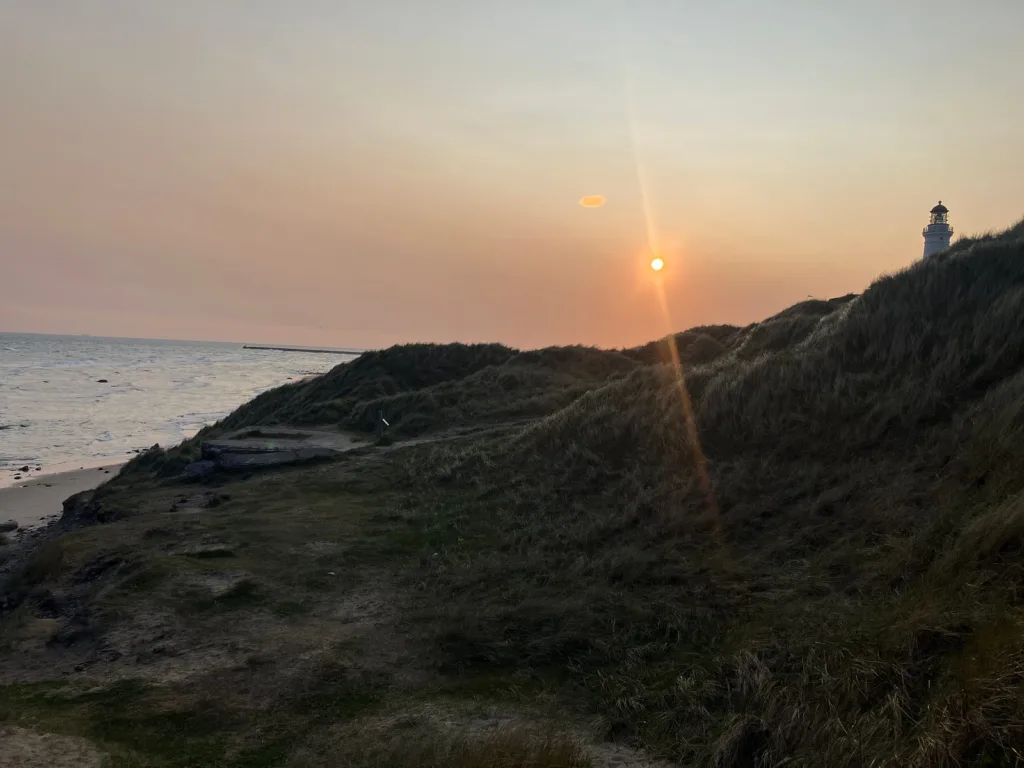 Hill overlooking sea with lighthouse and sun in background.