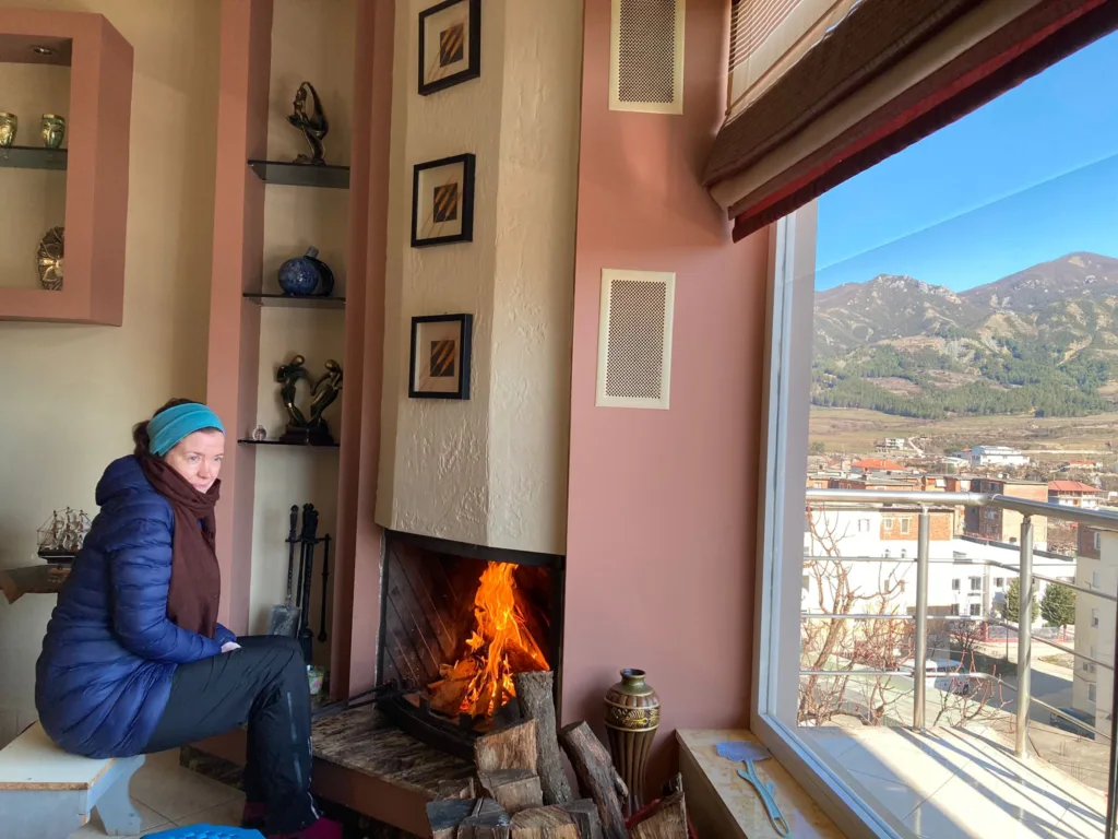 Person wrapped in puffy winter jacket and scarf sitting in front of a fire.