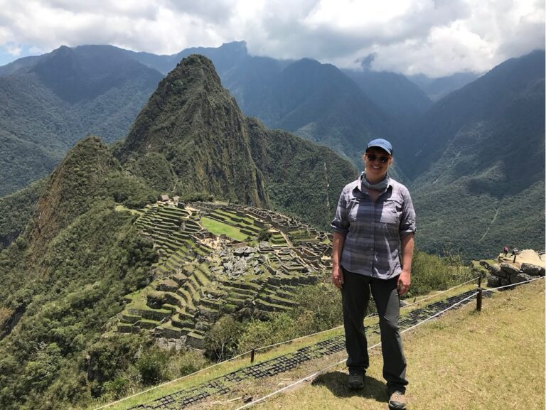 Nicole standing with blue cap, gray shirt, black pants. Background with green mountains and Machu Picchu.