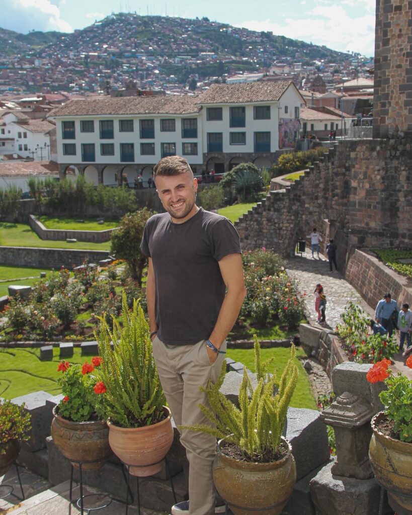 Man standing in black t shirt and khaki pants on stone stairway overlooking gardens in background.