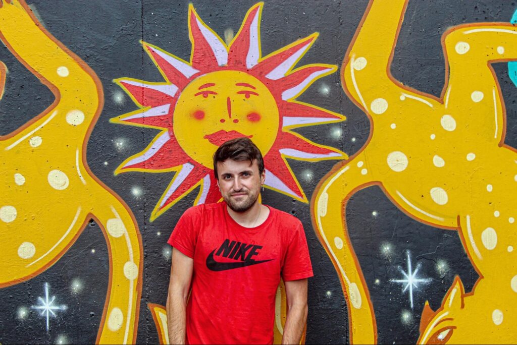 Man with brown hair and red shirt standing with hands in pockets in front of orange, blue, and yellow mural.