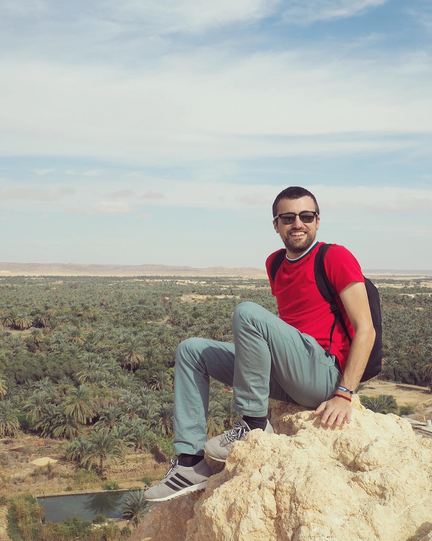 Man sitting in red shirt and jeans and sunglasses on a rock overlooking trees in background
