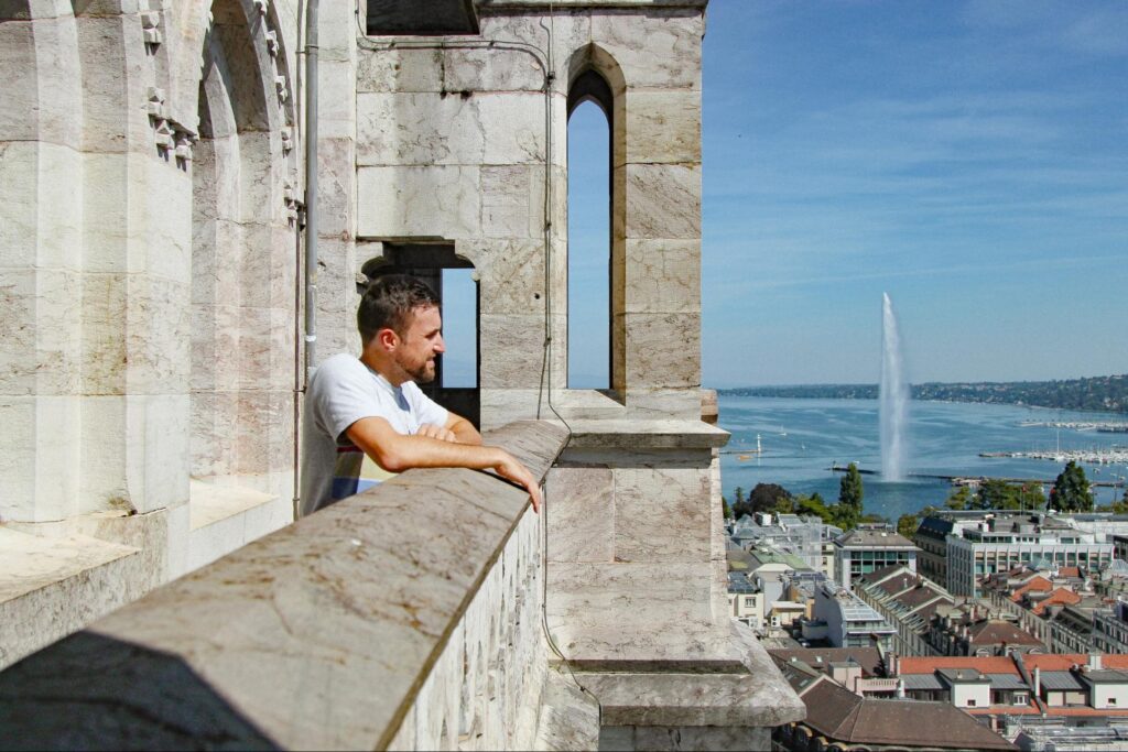 Man standing in white t shirt and looking over stone balcony over city with red roofs and large fountain and lake or seaside